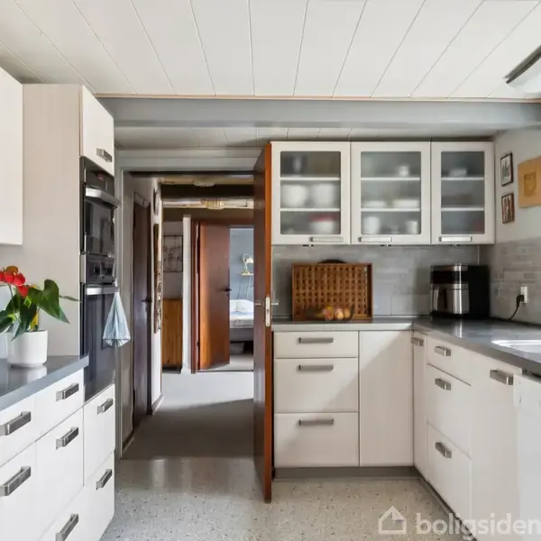 Køkken med hvide skabe, moderne apparater og grå bordplader. En køleskab med magneter og en plante på toppen. Et vindue til højre slipper lys ind i rummet.