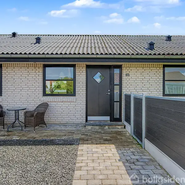 Et rækkehus med muret facade har en sort dør og to vinduer. Foran huset står to kurvestole og et bord på flisebelagt grund. Der ses en grusbelagt indkørsel.