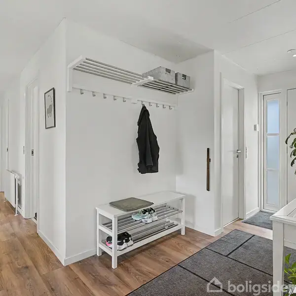Entréhall med skohylde og knagerække, jakke hænger; lyst rum med trælaminatgulv og to døråbninger. Indgang til højre med glasdør. Grøn plante står på hvid bænk.