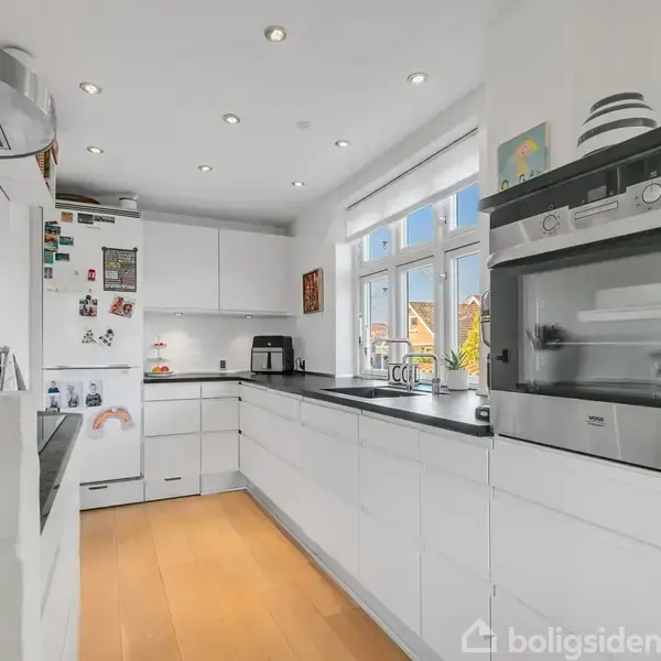 Et moderne køkken med hvide skabe og sorte bordplader. En ovn er indbygget i væggen, mens køleskab prydes med magneter. Vinduerne giver udsigt til et lysfyldt område.