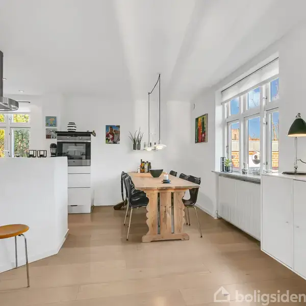 Spisebord af træ med seks stole står i et lyst køkken-alrum. På væggen hænger et ur. Bordet er dekoreret med lys og malerier ses i rummet.