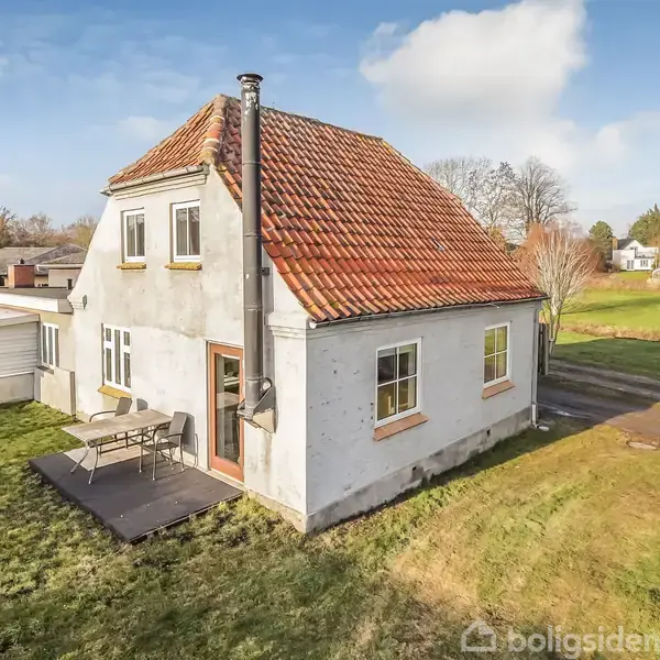 Et hvidt to-etagers hus med rødt tegltag står på en grøn græsplæne. En lille terrasse med bord og stole ved siden af huset. Omgivet af træer og flade landskaber.