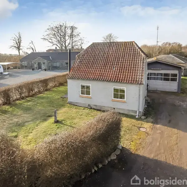 Hus med rødt tegltag og hvid facade, står stille ved siden af en garage. Omgivet af en græsplæne og hække i et villakvarter med flere bygninger i baggrunden.