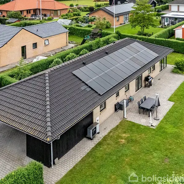 Et langt hus med solpaneler på taget ligger i et beboelseskvarter. En have og terrasse med bord og stole omgiver huset. Omkringliggende huse er synlige i baggrunden.