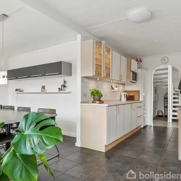 Køkken med lyse træskabe, udfører opbevaring, i et moderne hjem. Rummet har mørk flisegulv, spisebord til otte, grønne planter og en åben trappe. Hylder med dekorative figurer.