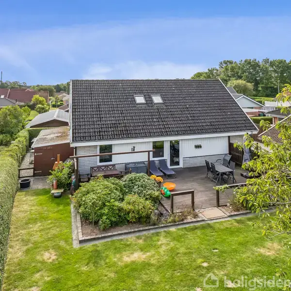 En hvid, enkelt-etagers villa har en terrasse med havemøbler og legeredskaber. Huset er omgivet af en velplejet have med buske og træer i et fredeligt boligområde.