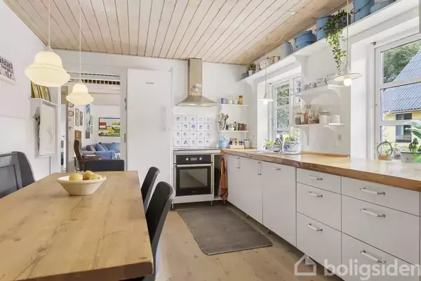 Et lyst køkken med træbord og sorte stole, en ovn med emhætte, hvidt skab og hylder med planter ved vinduerne, i et hjemligt miljø.