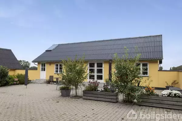 Et gult hus med mørkt tag står omgivet af blomsterkasser og planter. Foran huset er en stenterrasse med havemøbler placeret i en solrig have.