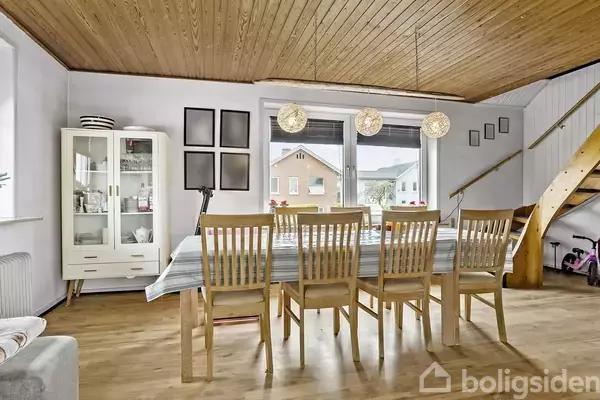 Spisebord omgivet af otte træstole står i et lyst rum med trægulv, træloft og store vinduer. En vitrineskab og en pyntet trappe ses i baggrunden.