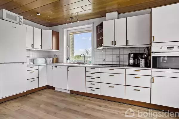 Et køkken med hvide skabe og skuffer er arrangeret langs væggen. En opvaskemaskine og køleskab integreres i designet. En kaffemaskine står på bordet ved siden af fliser. Udsigt til natur gennem vinduet.