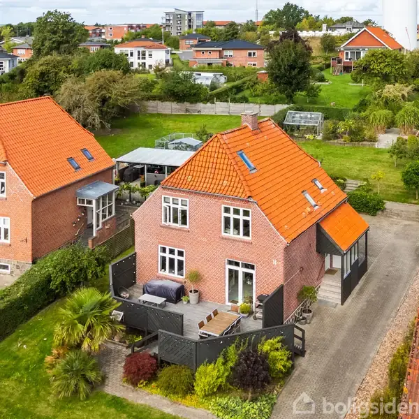 Hus med rødt tegltag og rød murstensfacade. Huset står roligt i et frodigt boligområde med grønne haver og indkørsler. Terrasse foran med sofaer og planter.