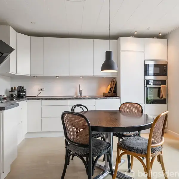 En rund spisebord med fire stole står i et moderne køkken med hvide skabe. En sort lampe hænger over bordet. Store vinduer og en reol med planter ses i baggrunden.