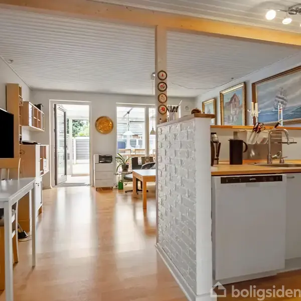 Køkken og stue i åben planløsning; køkkenbord med apparater; spisebord i stuen; store vinduer giver lys; udgang til terrasse. Skandinavisk indretning med trægulve og lyse farver.