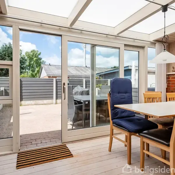 Spisebord med træstole står på en lys veranda. En glasdør åbner til en terrasse med fliser, omgivet af hegn. Murstensvæg bagved har hængende lampe og landskabsmaleri.