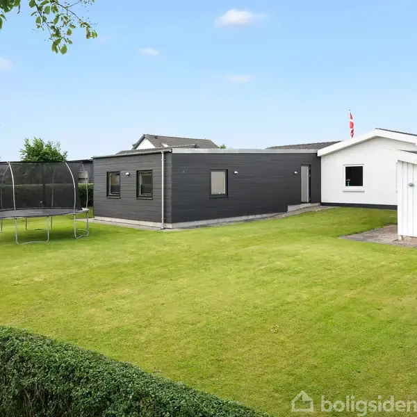 En grøn græsplæne prydet med en trampolin i forhaven af et moderne hus med fladt tag. Hækken omgiver plænen, og solen skinner i den blå himmel.