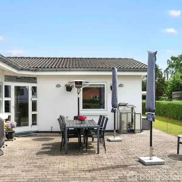 En udendørs terrasse med sorte havemøbler står på grå fliser foran et hvidt hus. Bordet har en rød blomsterdekoration. Der er parasoller, en grill og en lille bålplads.