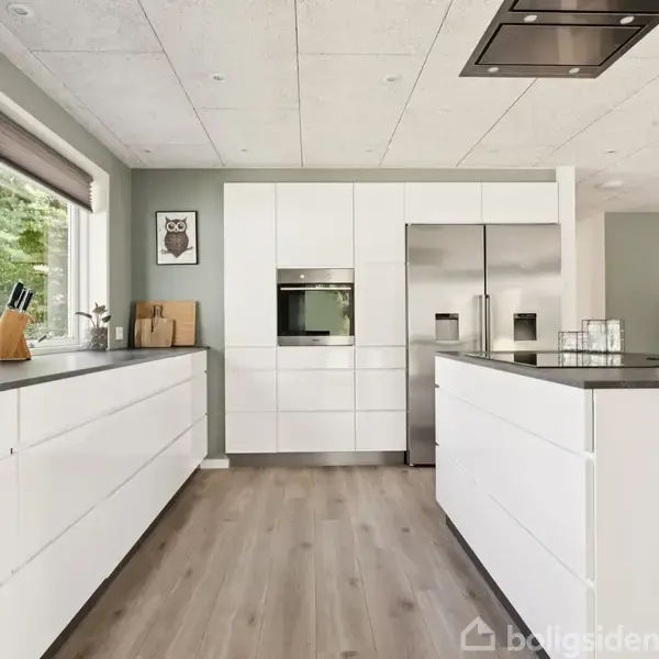 Hvidt køkken med moderne skabsmoduler, integreret ovn og køleskab. Køkkenø står centralt. Store vinduer giver naturligt lys. Trægulv og grå vægge skaber en lys og åben atmosfære.