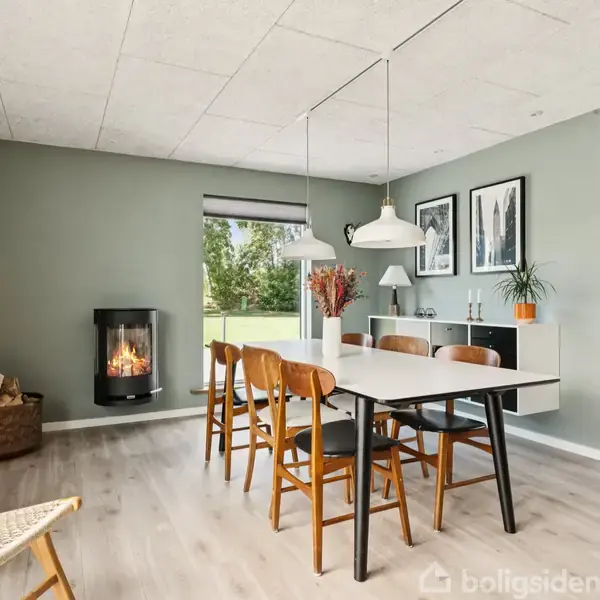 Spisebord med træstole står i en moderne spisestue med brændeovn på væggen. Grønne vægge, hylder med planter og udsigt til have gennem vinduet skaber et hyggeligt miljø.