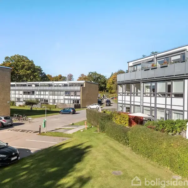 Flere etagebygninger står stille omkranset af græsplæner og parkeringspladser med parkerede biler. Solen skinner, og træer ses i baggrunden.