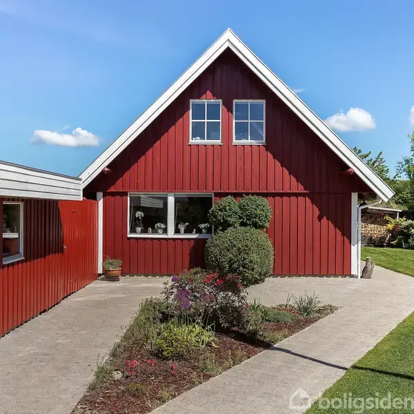Rødt træhus står stille, omkranset af velholdt have med blomsterbede og grøn græsplæne under en blå himmel. Huset har to vinduer på taget og et stort vindue i stueetagen.