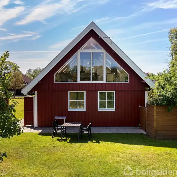Et rødt hus med hvide kanter står i en grøn have; foran er et havemøblement af bord og stole. Huset har store vinduer øverst i gavlen, omgivet af træer og græsplæne.