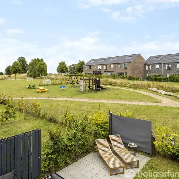 To solstole står på en flisebelagt terrasse med udsigt over en legeplads med gyngestativ. Rækkehuse ligger i baggrunden omgivet af grønne områder og himlen er klar.