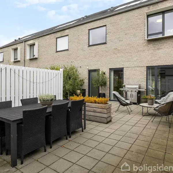 Sort havebord med stole står på en flisebelagt terrasse ved en bolig. To stole og en grill er placeret nær en murstensvæg. Hegn og planter omkranser terrassen.