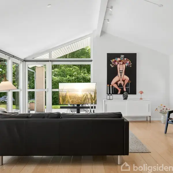 Sort sofa står foran et stort vindue med udsigt til en grøn have. En lampe står ved siden af sofaen, og et farverigt billede hænger på væggen. En fjernsynsskærm viser naturbilleder.