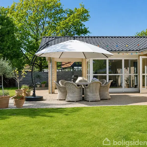 En terrasse med en rund spisebord og stole under en stor parasol. Terrasse omgivet af frodig have med potteplanter; huset er set i baggrunden med store vinduer.