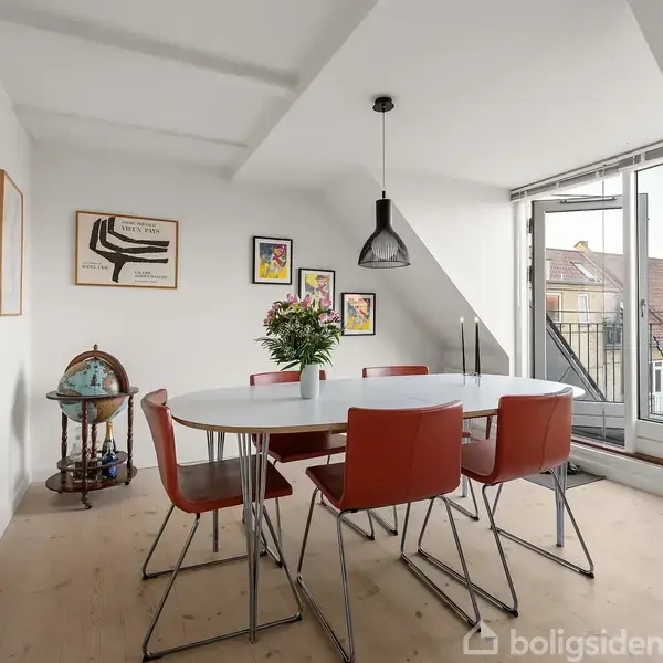 Et rundt bord med røde stole står i spisestuen. En lampe hænger over bordet, og rummet har kunst på væggene. Store vinduer fører til en altan med udsigt over tagene.