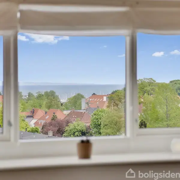 Et vindue giver udsigt over en by med røde tage og frodige træer. I forgrunden står små dekorationer, herunder en plante, på vindueskarmen.