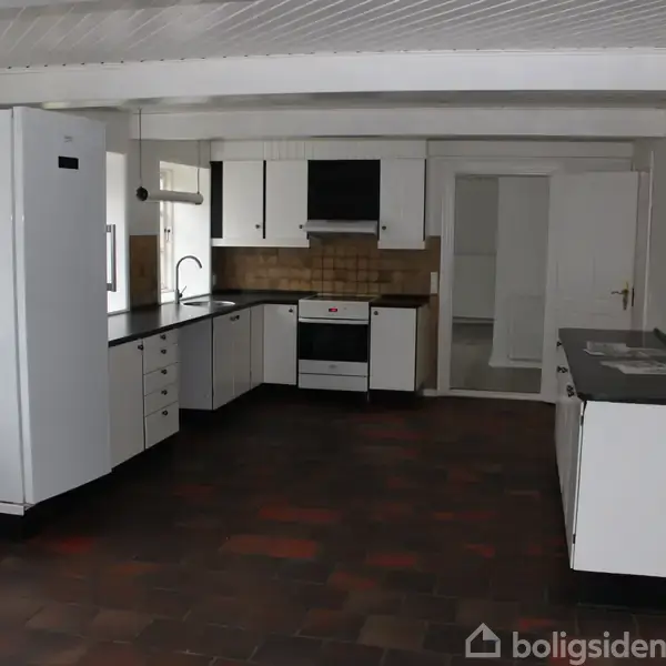 Et køkken med hvide skabe og sorte bordplader. Der er et komfur, en ovn og en stor hvid køleskab. Rummet har hvide vægge og et mørkt flisegulv.