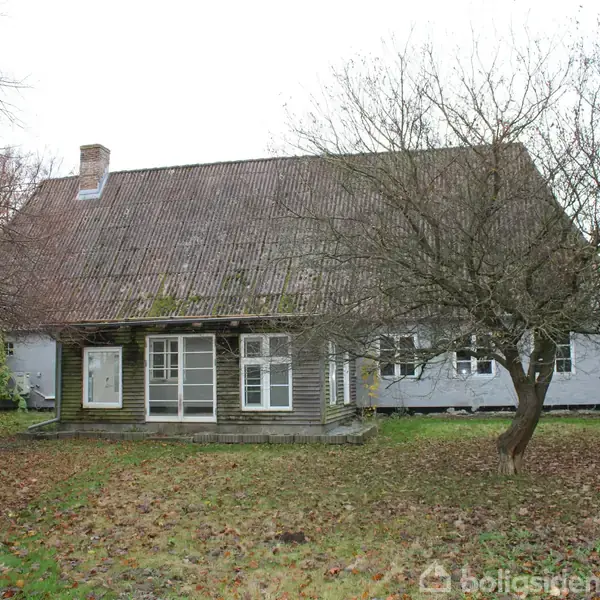 Et gammelt, træhus står stille omgivet af en græsplæne med visne blade og nøgne træer. Den store tagryg og skorstenen stiger op under den grå, overskyede himmel.