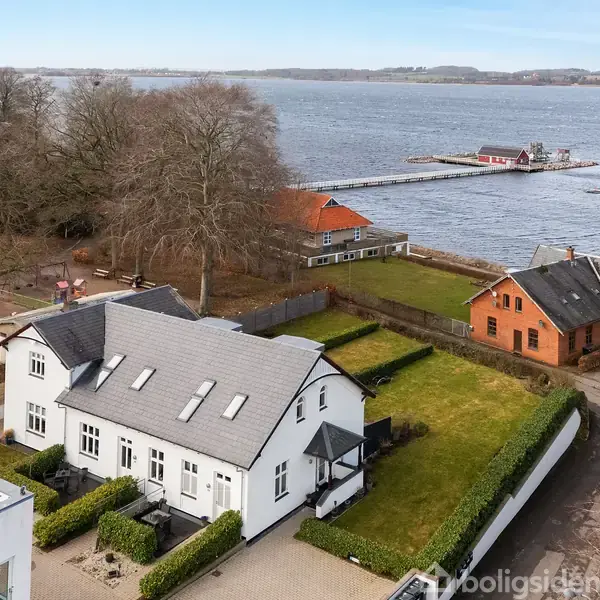 Hvidt hus med grå tagsten står ved siden af en velplejet have, tæt på en sø. Baggrunden viser en bådebro og flere huse langs bredden med store træer omkring.