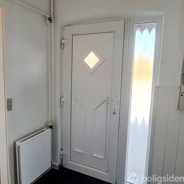Hvid dør med diamantformet vindue står i en lys gang ved siden af en radiator. En smal, frostet rude til højre slipper lys ind. Der er en eltavle på væggen.
