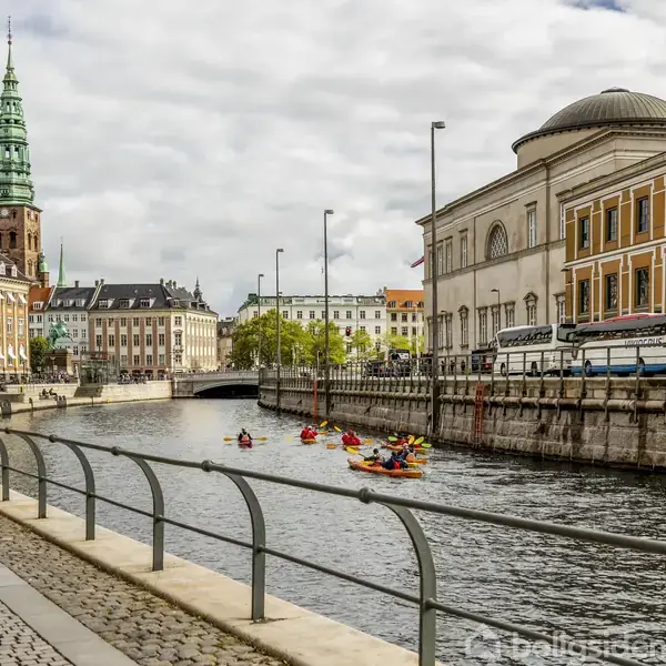 Flere farverige kajakker sejler på en kanal omgivet af gamle bygninger med en kirke med et grønt spir i baggrunden. Brostensbelagt promenade langs kanalen.