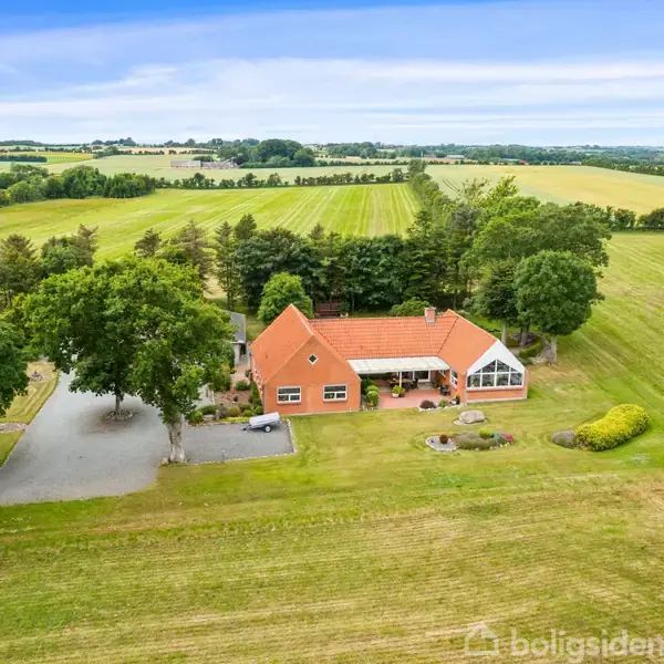 Et rødt murstenshus med orange tag omgivet af grønne marker og store træer. Grusvej fører til huset, der har en anlagt have. Landskabet er landligt og åbent.