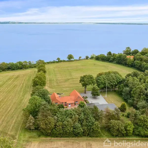 Et rødt teglhus omgivet af træer står i en stor, åben mark ved en blå sø. Baghaven er græsklædt, og der er skov på den ene side.