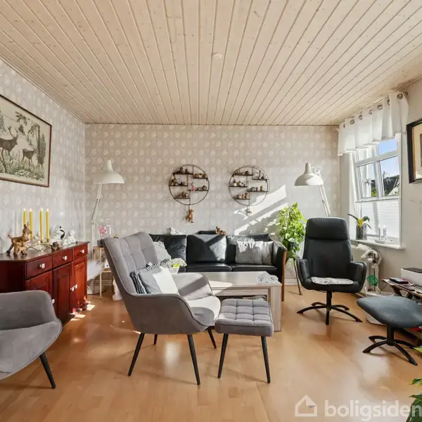 Stue med møbler som sofa, stole og et sofabord, omgivet af lyse tapetvægge og træloft. Vinduerne lader sollys strømme ind, og der er planter samt dekorative billeder og lamper.