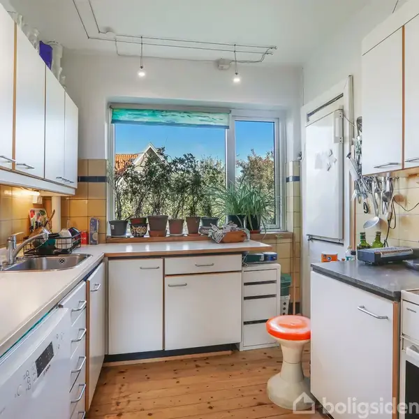 Køkken med hvide skabe og moderne apparater, herunder opvaskemaskine og ovn. En orange skammel står ved sort bordplade. Vindue viser udsigt til grønt buskads udenfor.