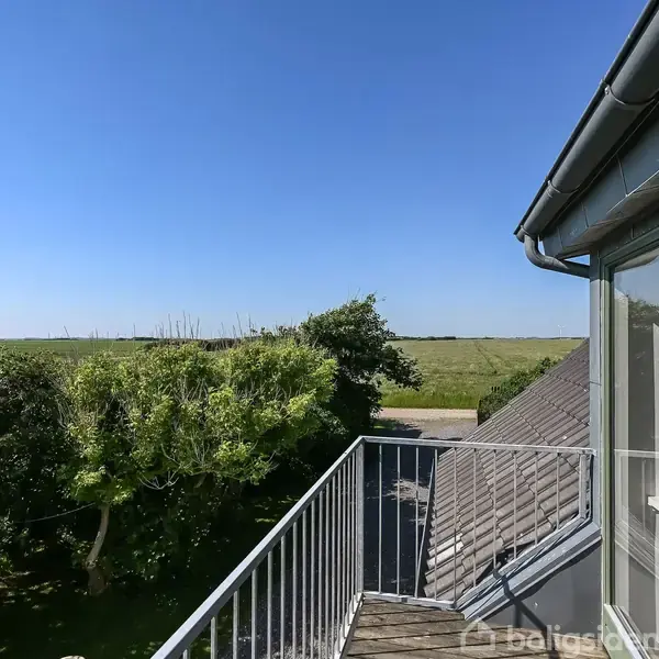 Balkon med hvidt rækværk fører ud fra husets vinduer, hvor træer og grønne marker strækker sig under en klar blå himmel.