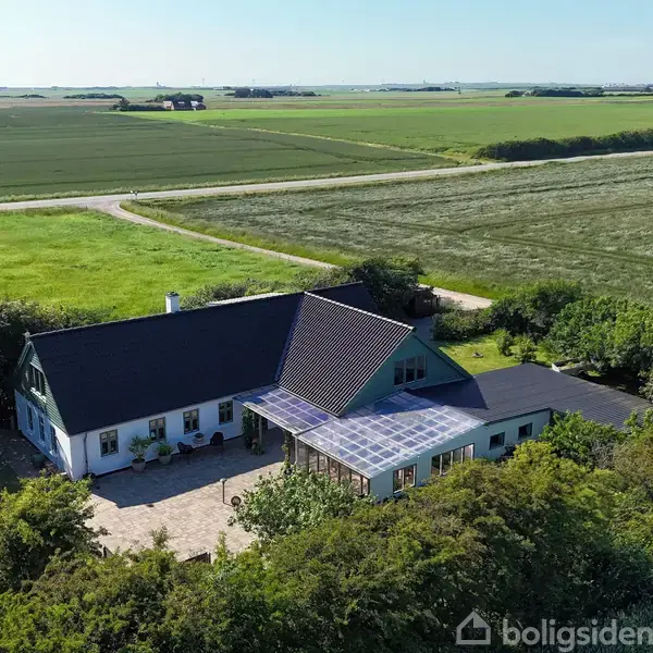 Hus omkranset af frodige marker, har et mørkt tag og solcellepaneler. En stenbelagt gårdsplads foran, omgivet af grønne buske og træer. Klar blå himmel.