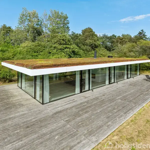 Et moderne hus med glasvægge har et grønt tag og står på en stor træterrasse. Det er omgivet af frodige træer og buske i en naturskøn have.