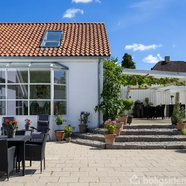 Et moderne køkkenalrum med hvidt interiør har et sort køkkenbord, hængende lamper og et rundt spisebord med stole. Store vinduer giver udsigt til en terrasse og have.