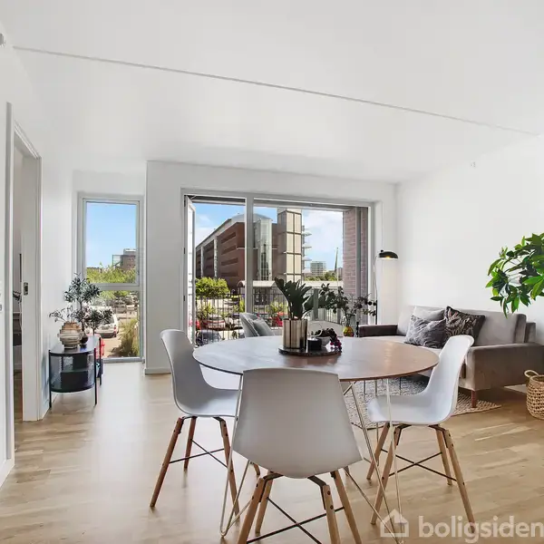 Et rundt spisebord med moderne stole står i en lys stue. En stor plante og sofa ligger ved siden af, mens glasdøre fører til en altan med udsigt over byen.