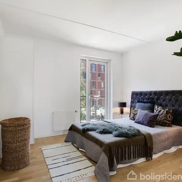 Soveværelse med en dobbeltseng med mørkt hovedgærde, opredt med tæpper og puder. Ved siden af står en plante og en gulvlampe. Et stort vindue giver lysindfald.
