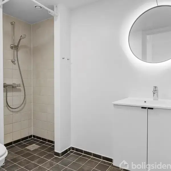 Badeværelse med brusekabine til venstre, hvidt toilet og moderne vaskenhed med rund oplyst spejl til højre. Fliser på gulvet og lys indretning giver et rent udtryk.