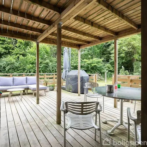 Terrasse med trægulv, indeholder et rundt glasbord med fire stole, en grill og en sofa. Overdækket af trætag og omgivet af træer og en bygning med røde vinduer.