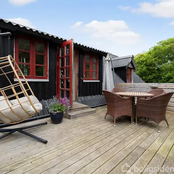 Gyngestol i rattan står på en træterrasse ved et hus med sorte vægge og røde vinduer. Et rundt bord med to stole ligger tættere på. Grønne træer i baggrunden.