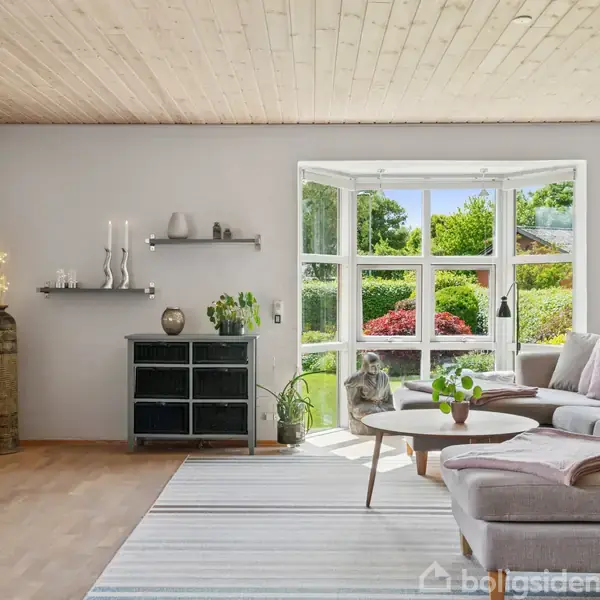 Sofa står ved store vinduer, som åbner ud til en grøn have. Indretningen inkluderer hylder med dekorative genstande og et lille bord med blomster.